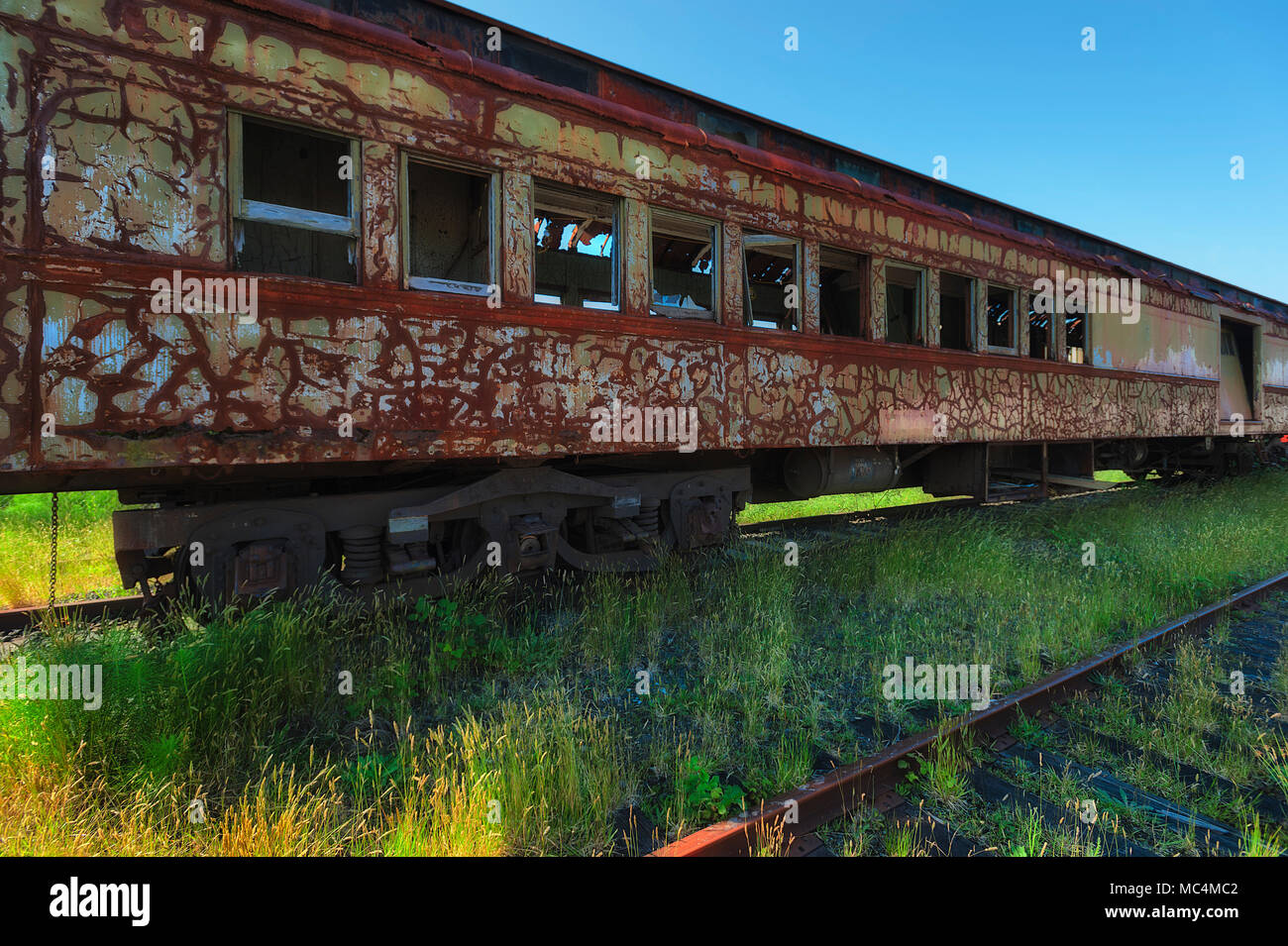 A rusted abandoned passenger rail car sits on likewise train tracks surrounded by weeds and grass. Stock Photo