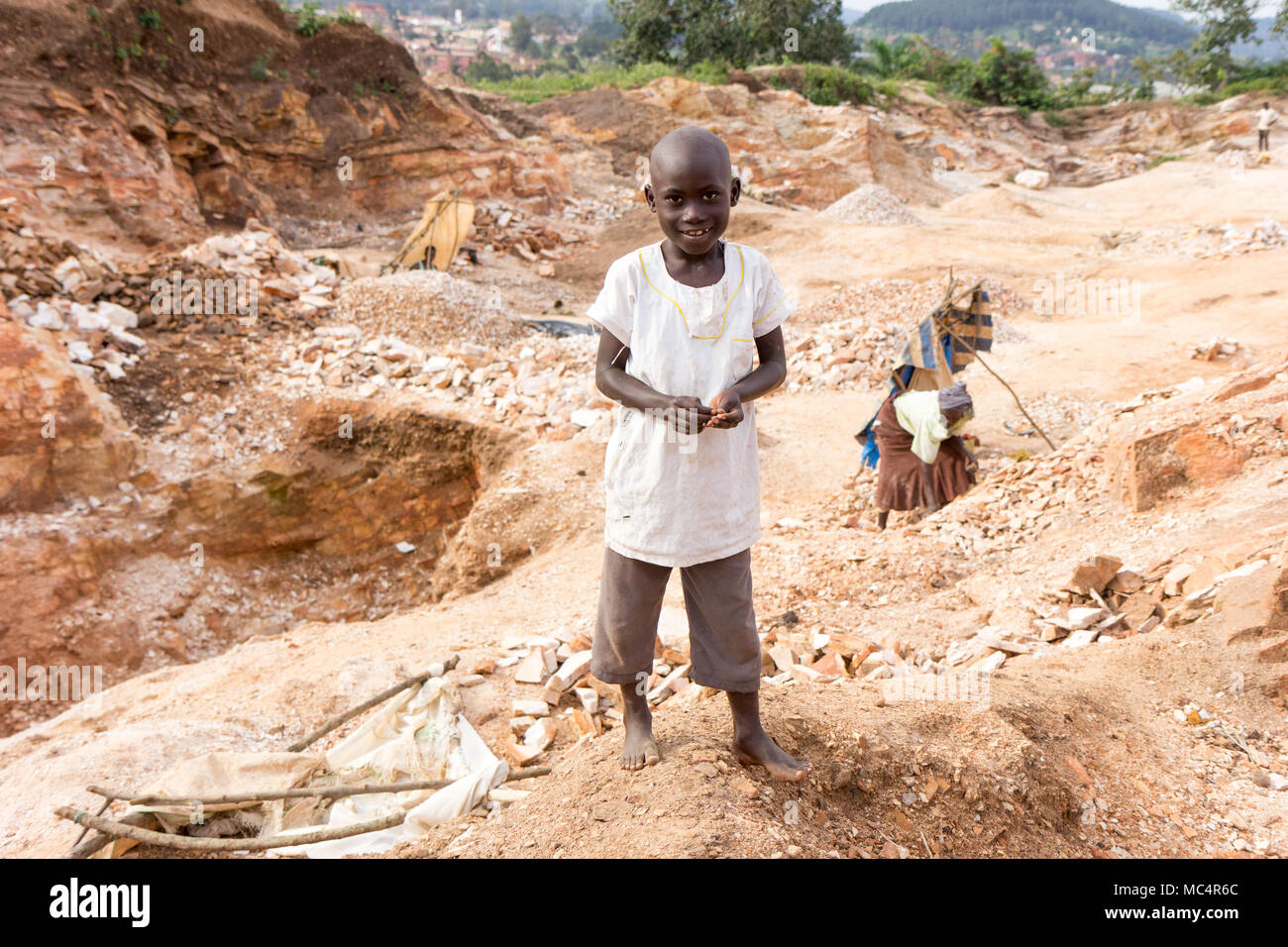Lugazi, Uganda. June 18 2017. A smiling Ugandan boy standing in a quarry. People in the background are breaking rocks into small slabs for sale to the Stock Photo