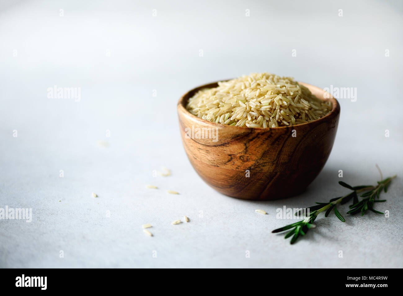 Raw organic brown rice in wooden bowl and rosemary on light concrete background. Food ingredients. Copy space Stock Photo