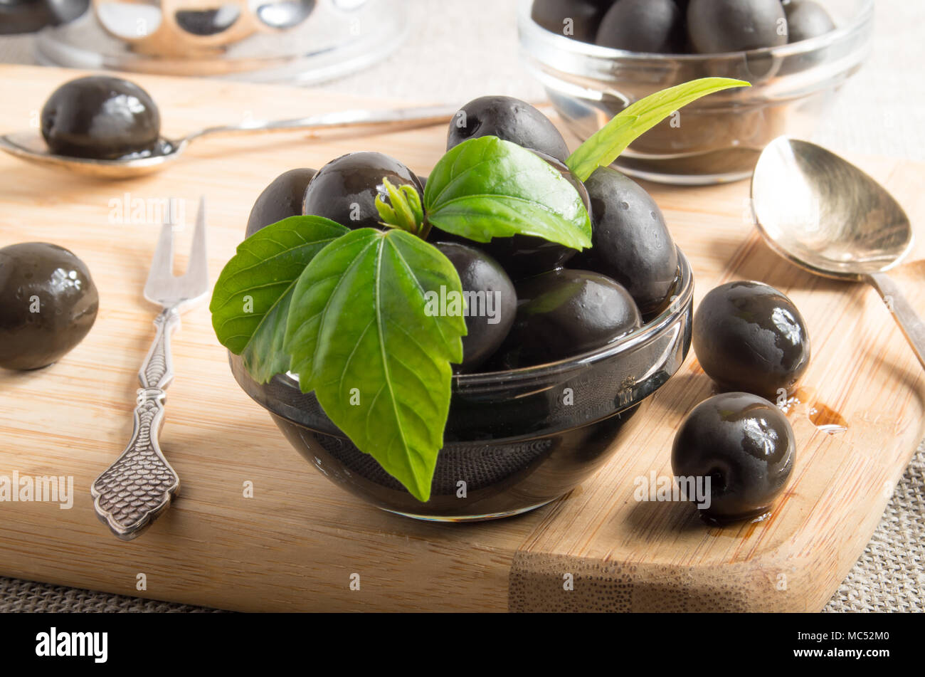 Black olives in glass cups decorated with green leaves on a wooden tray and vintage crockery with shallow depth of focus Stock Photo