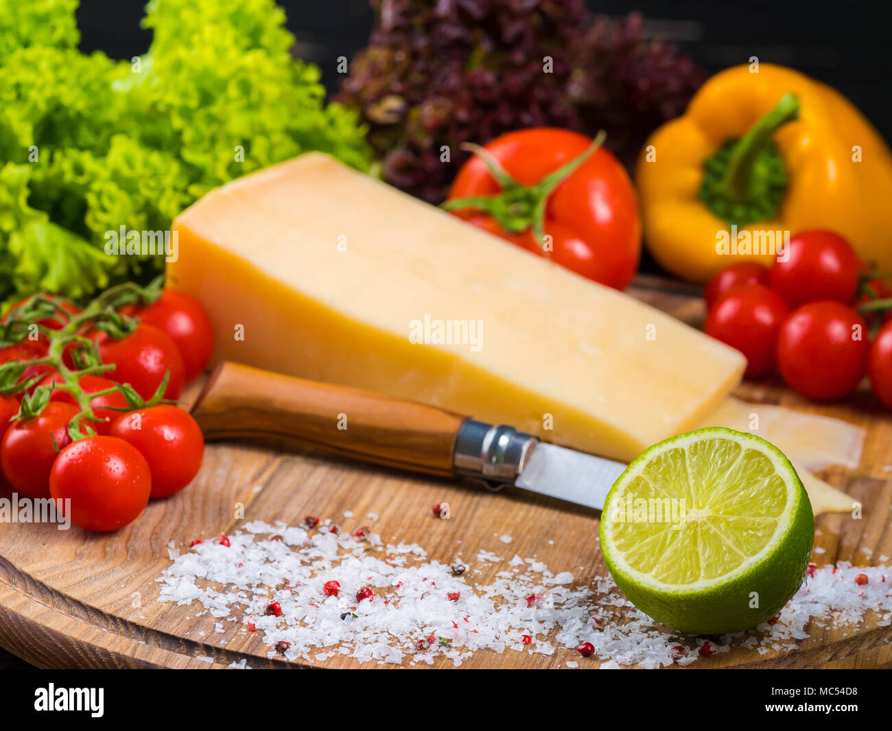 wooden board with parmesan cheese, fresh cherry tomatoes, lettuce, sea salt, lime and red pepper. The knife for cutting vegetables. Appetizing still l Stock Photo