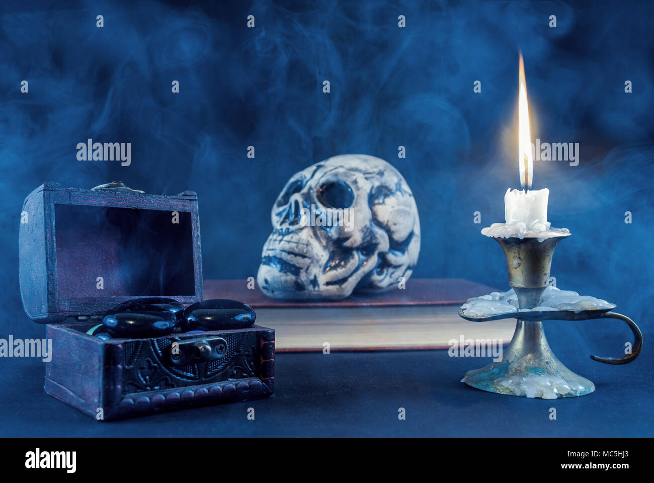 Mystical box with black rock and candle in candlestick burning and skull on book blurry in background, smoked and dark scene. Halloween concept. Stock Photo