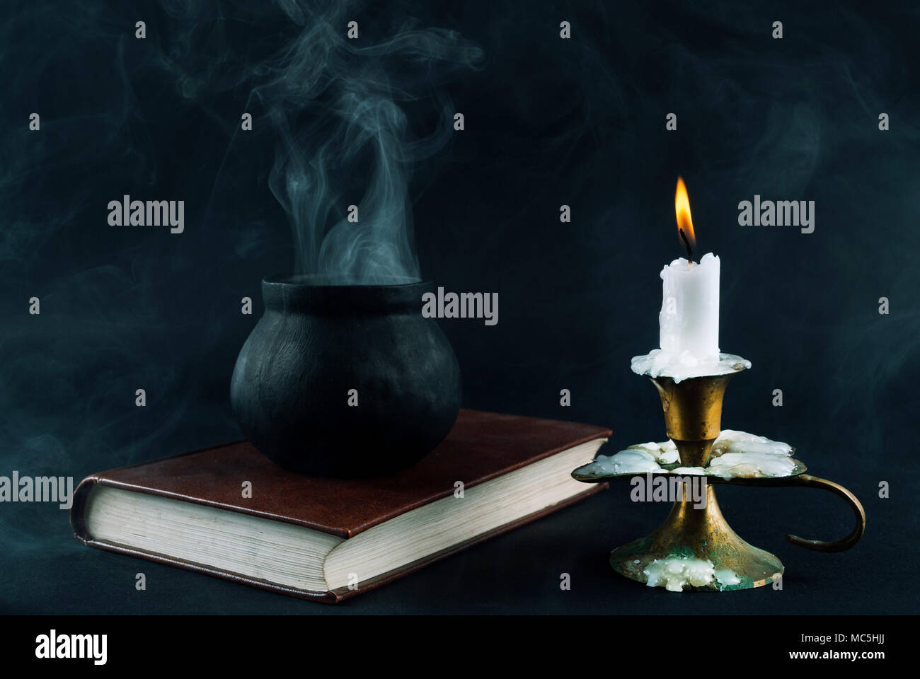 Wicker magic pot is smoked on the old book next to candle in old candlestick black and smoked background. Halloween concept Stock Photo