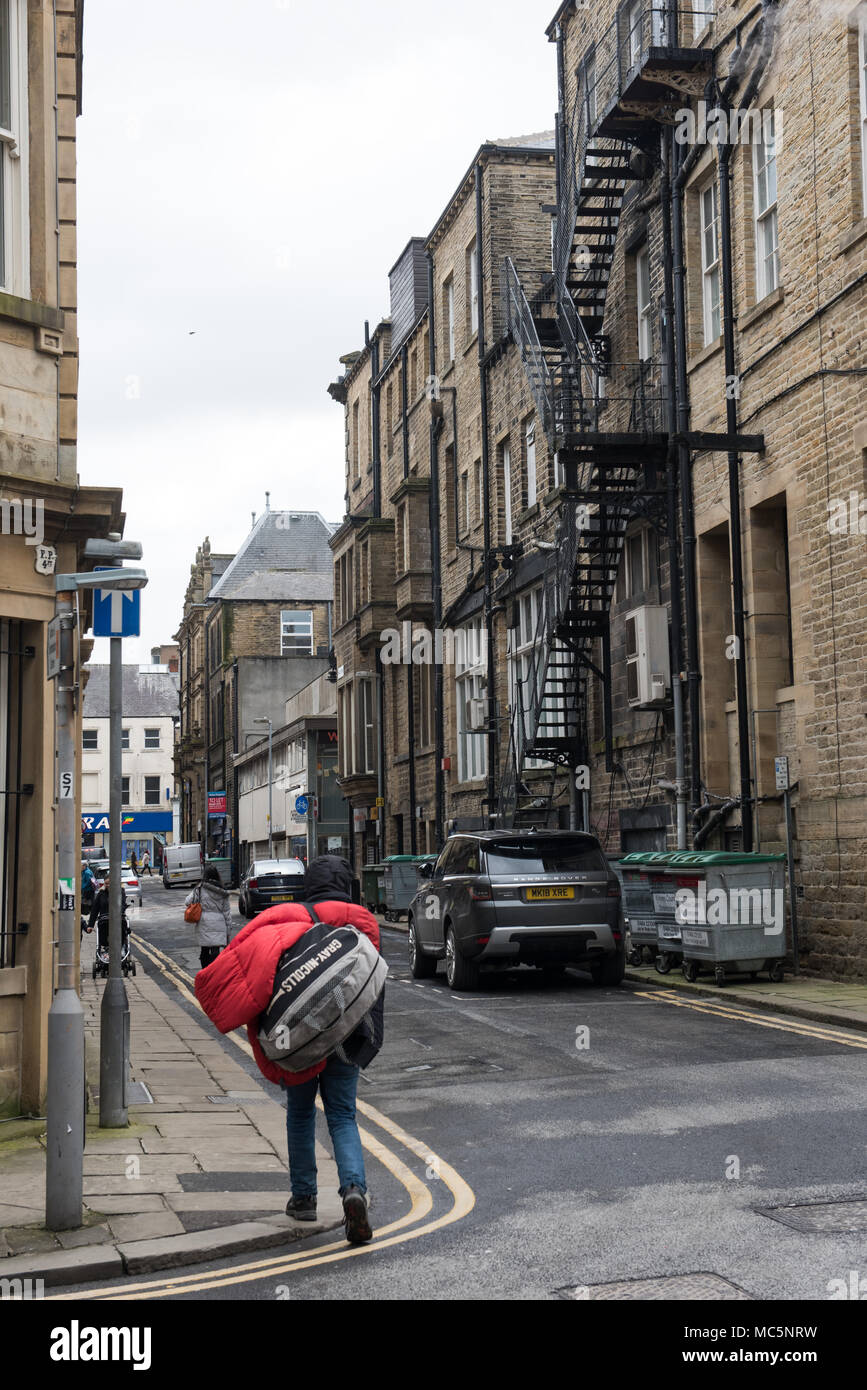 A person with a large red duffle bag walks down a narrow backstreet lined with stone buildings, bins, and a metal fire escape in a British town Stock Photo