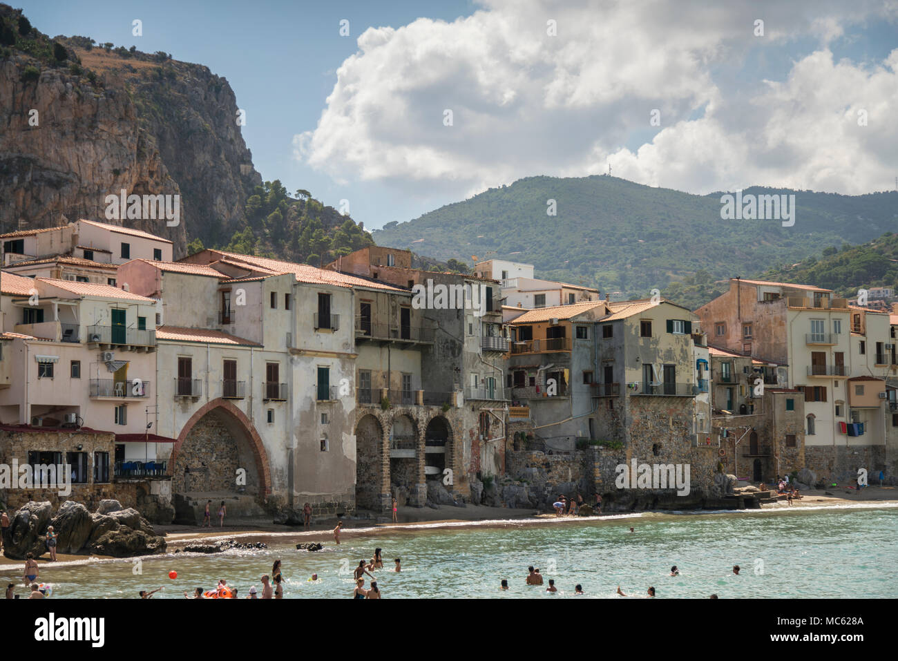 Rocky headland and historic buildings overlooking the sandy beach at Cefalu, Sicily, Italy, with holidaymakers swimming in the sea. Stock Photo