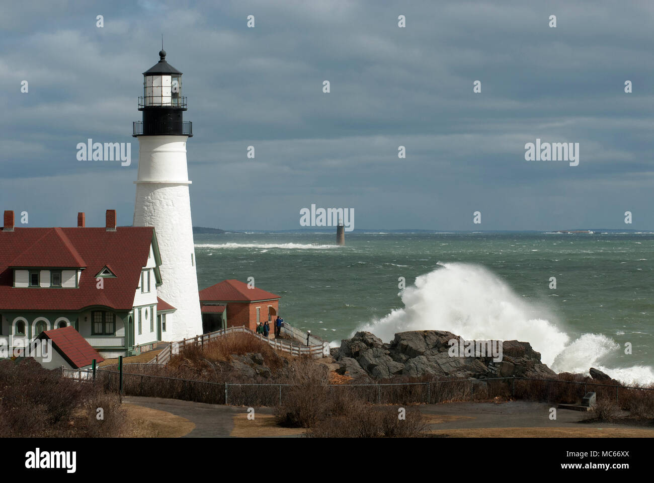 Giant waves crash over rocks by Portland Head lighthouse and out at Ram Island Ledge light as sun breaks through sotrm clouds in Maine. Stock Photo
