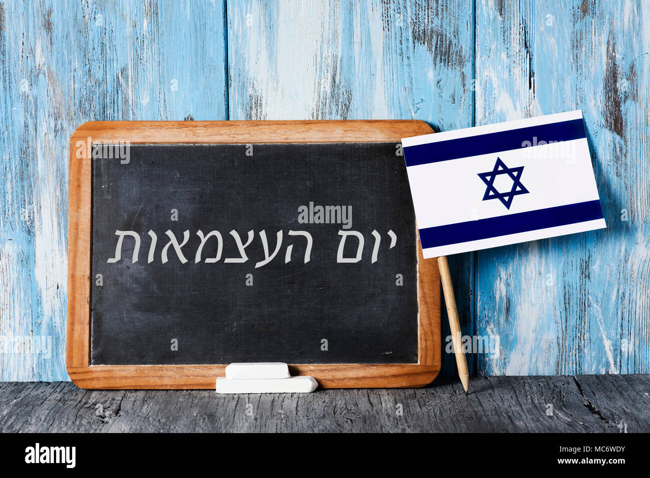 a wooden-framed chalkboard with the text independence day written in hebrew in it, some pieces of chalk and an israeli flag on a rustic wooden surface Stock Photo