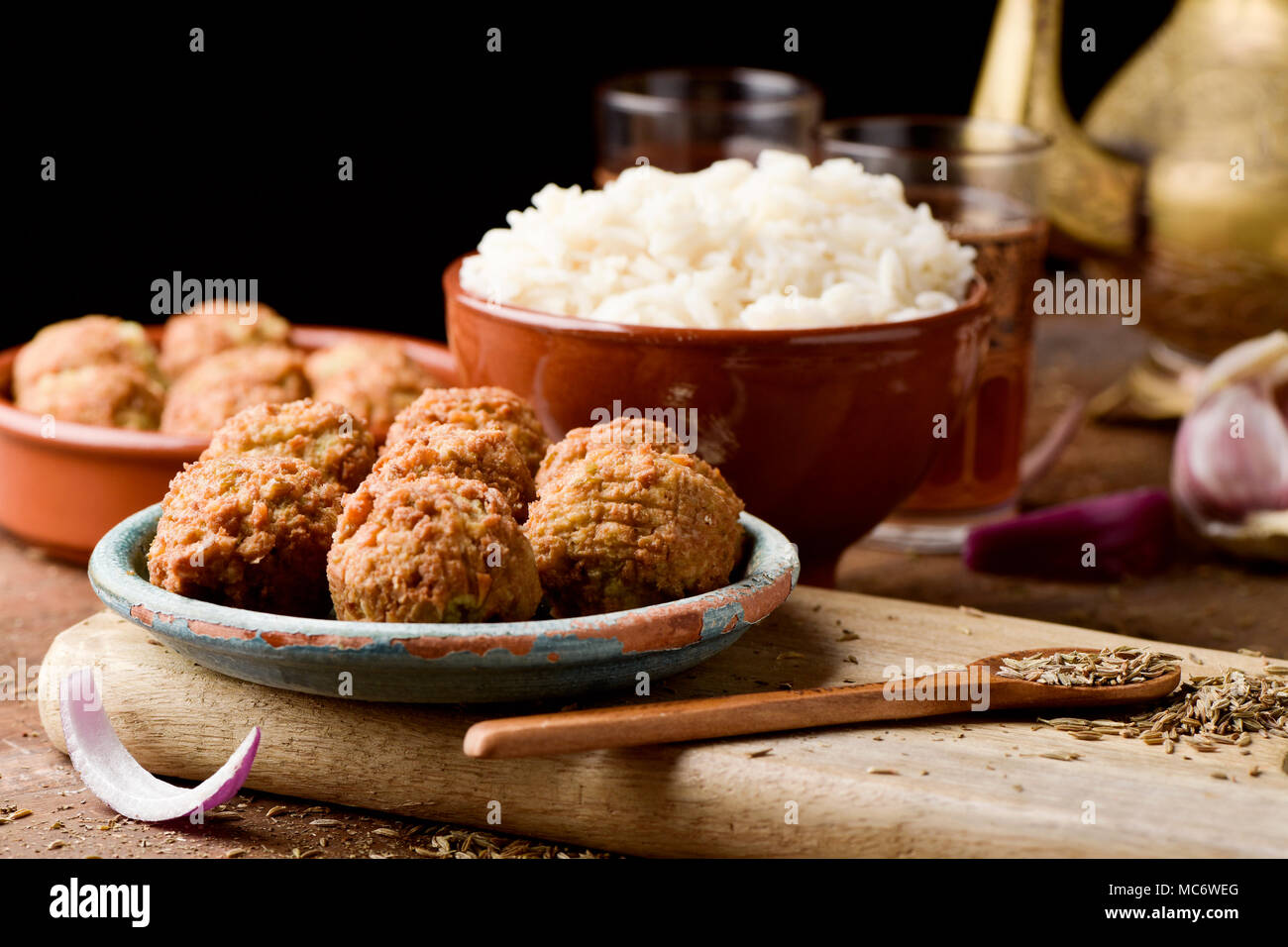 some falafel in a green earthenware plate and a brown earthenware bowl with long grain rice on a rustic wooden table, with a golden teapot and some or Stock Photo
