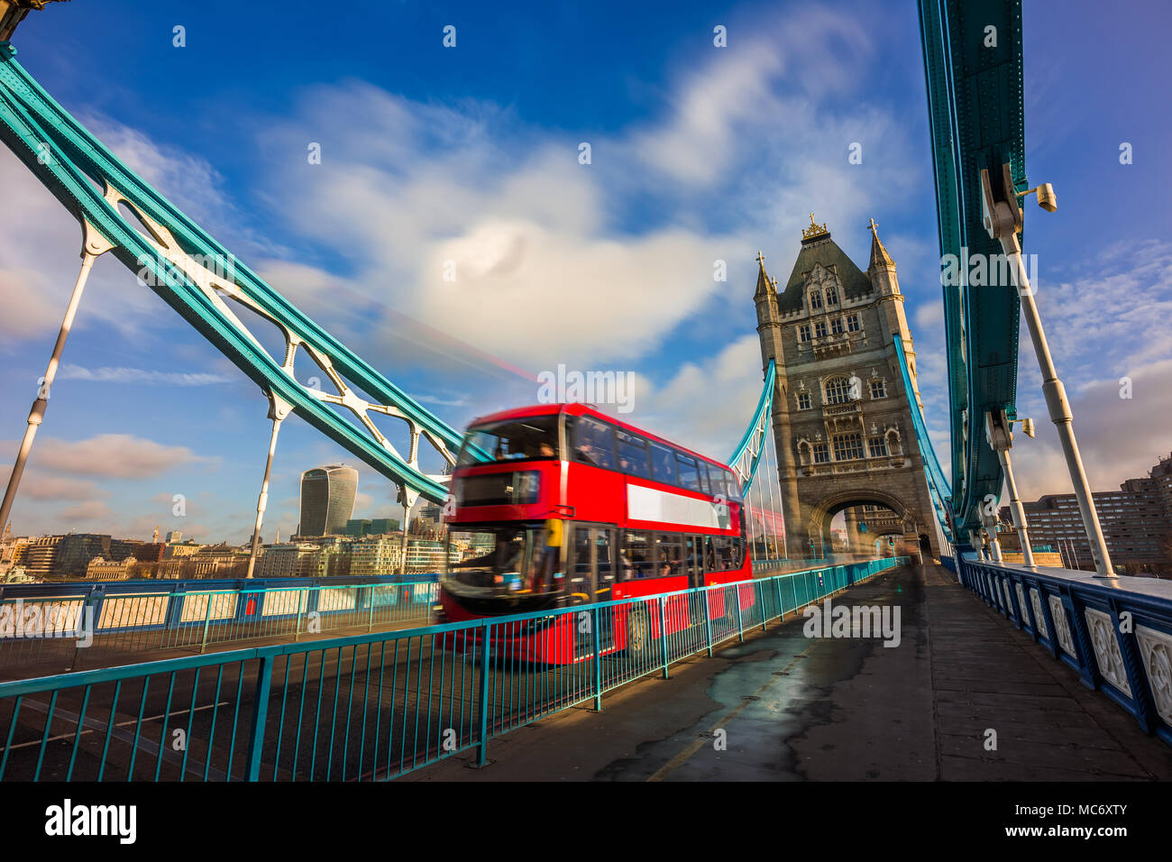 London, England - Iconic red double-decker bus in motion on famous Tower Bridge with skyscraper of Bank District at background. Blue sky and clouds Stock Photo
