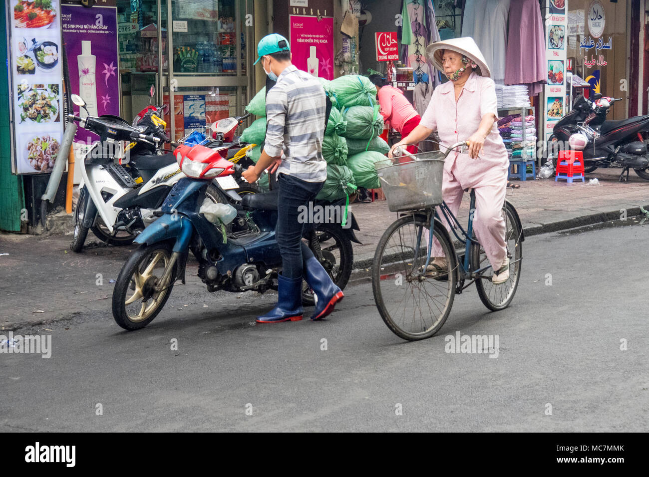 An elderly woman wearing a pink outfit and a conical hat cycling on a street in Ho Chi Minh City, Vietnam. Stock Photo