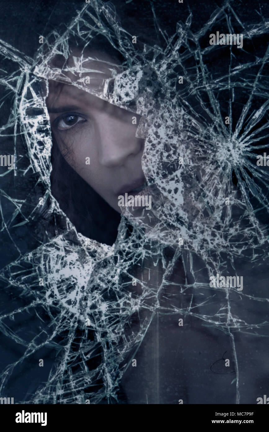 Closeup of a young caucasian woman face looking from behind a broken window glass, conceptual dramatic portrait. Stock Photo