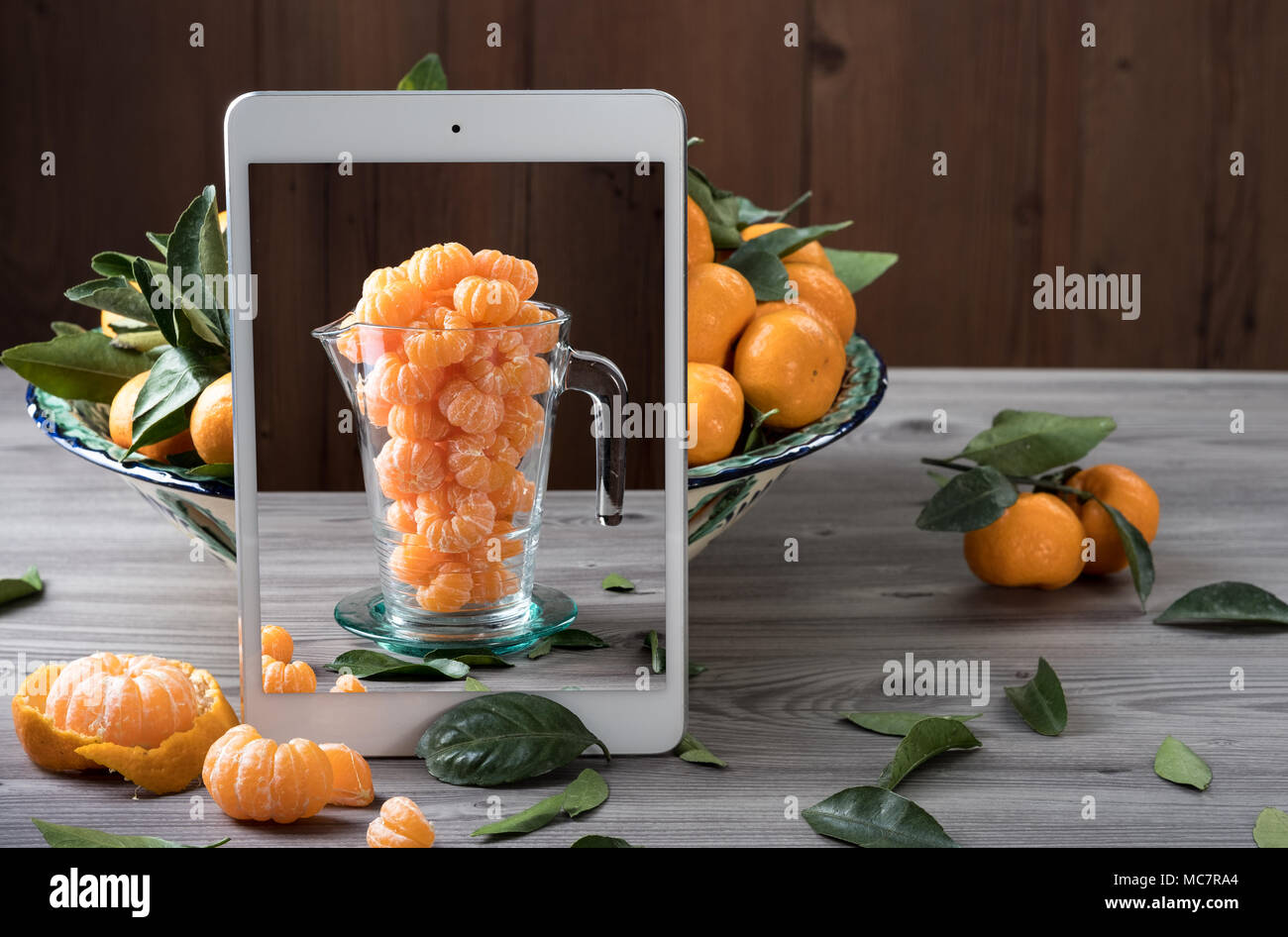 White mobile tablet standing in front of ceramic dish filled fresh tangerines on gray wooden table. Pop up visual effect. Creative food composition. Stock Photo