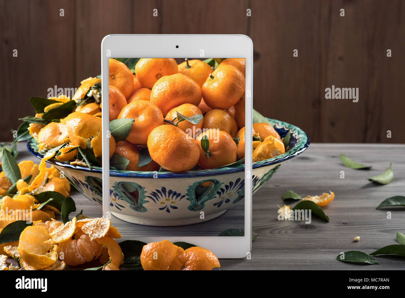 White mobile tablet standing in front of ceramic dish filled fresh tangerines on gray wooden table. Pop up visual effect. Creative food composition. Stock Photo