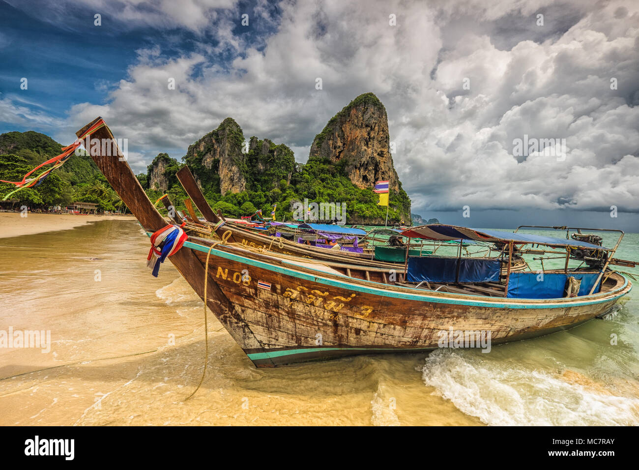 Colorful traditional longtail Thai boats decorated with ribbons. Tropical sand beach with limestone cliff and cloudy sky on background. Thailand. Stock Photo