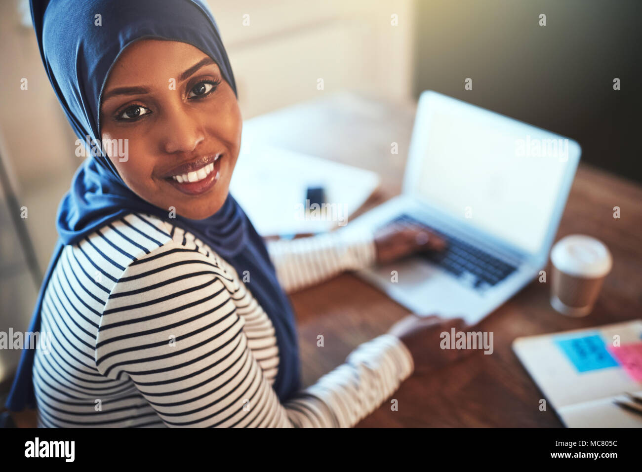 Smiling young Arabic female entrepreneur wearing a hijab sitting a desk ...