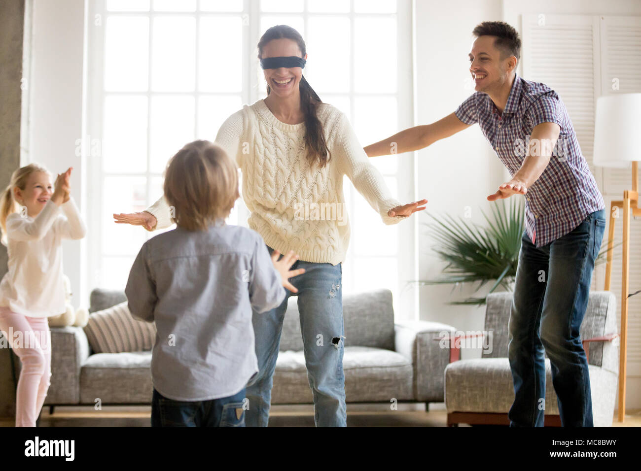 Family playing hide and seek game together with blindfolded mother, happy kids and parents having fun in living room, children hiding clapping hands e Stock Photo