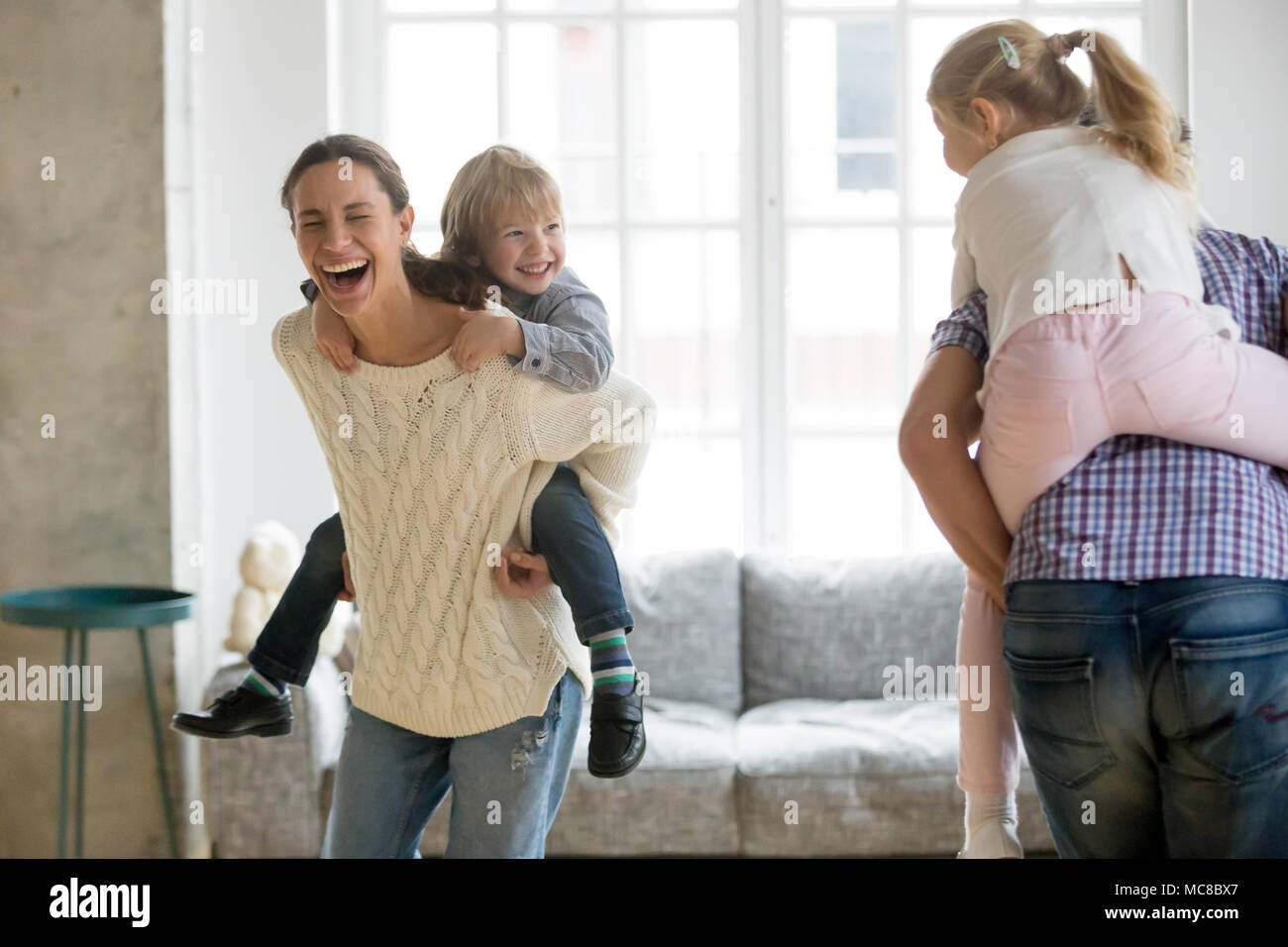 Happy mother laughing piggybacking little son playing with family at home, cheerful parents carrying kids on back having fun together, children boy an Stock Photo