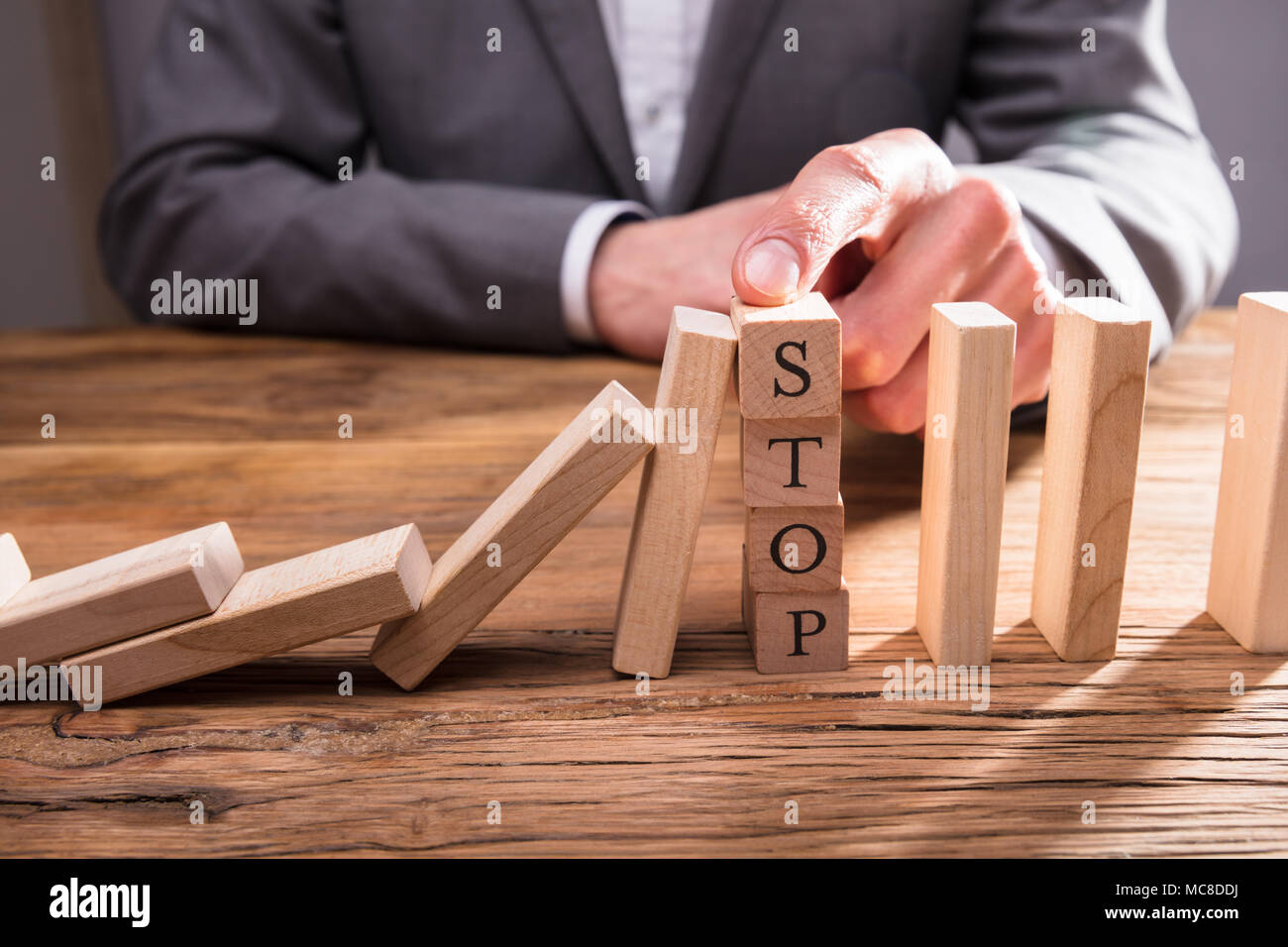 Businessperson's Finger Over Wooden Blocks With Stop Text Stopping Falling Dominos On Wooden Desk Stock Photo