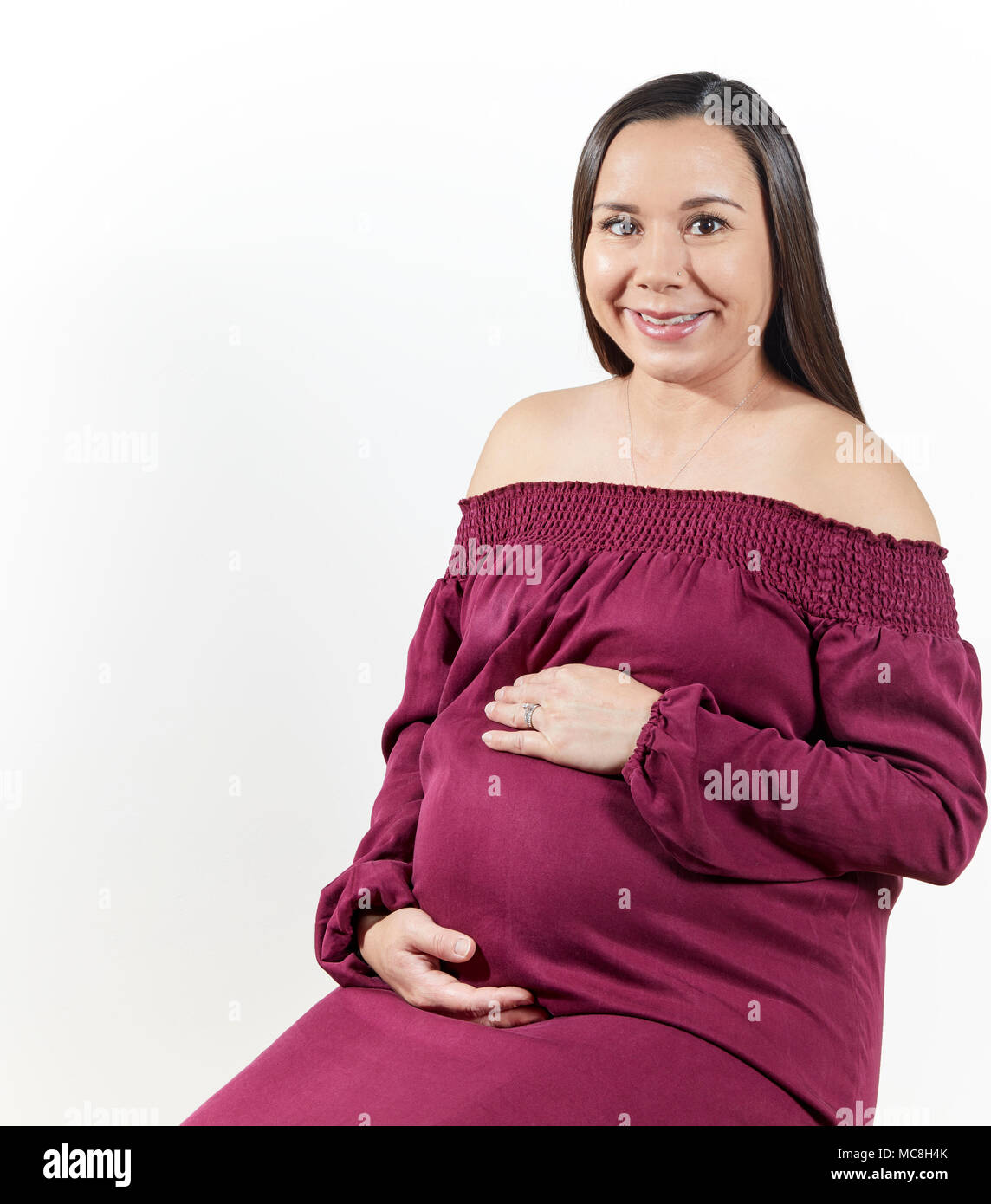 Attractive pregnant woman with her hands on her tummy sitting down isolated on white Stock Photo