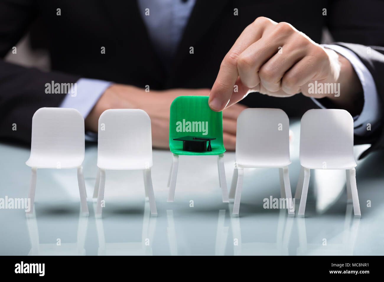 Close-up Of A Businessperson's Hand Choosing Green Chair With Graduation Hat Stock Photo