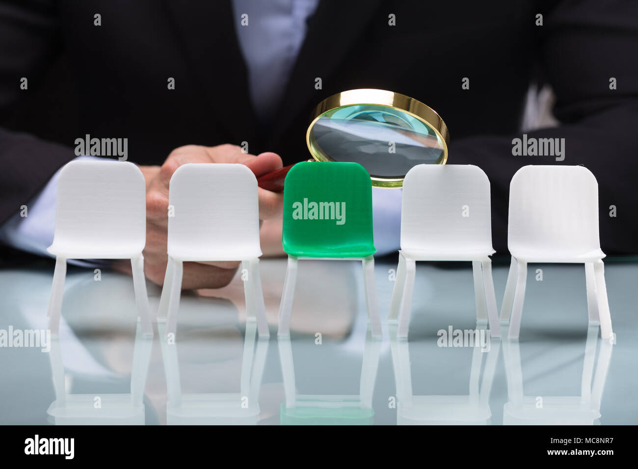 Close-up Of A Businessperson's Hand Looking At Vacant Chairs Through Magnifying Glass Stock Photo