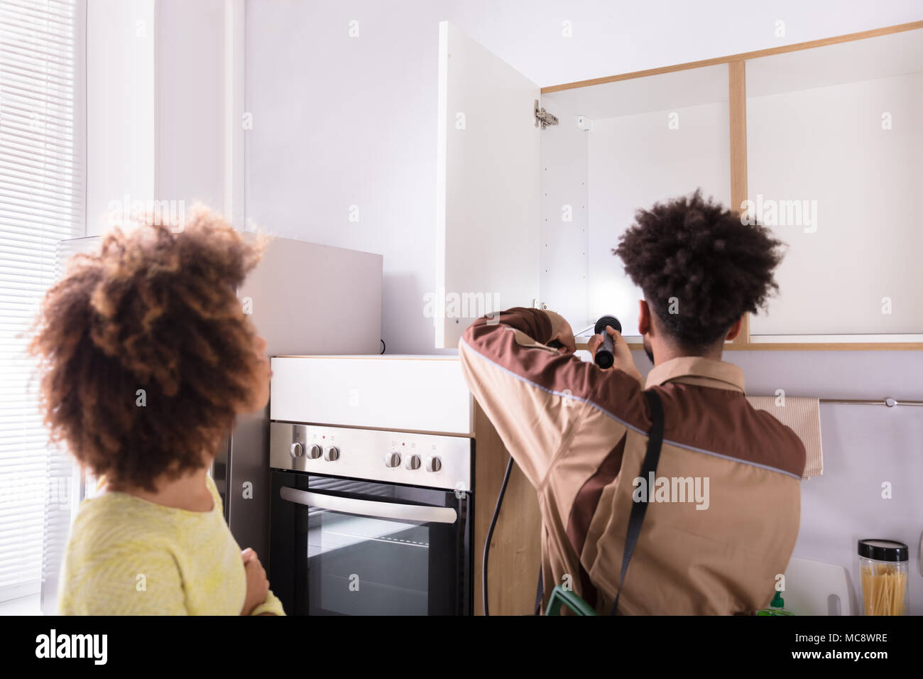 Woman Looking At Pest Control Worker Spraying Insecticide On Shelf Of Domestic Kitchen Stock Photo
