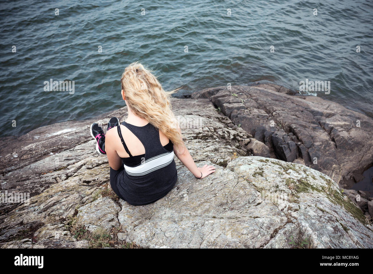 Rear view of a blond young woman sitting on a rock looking at view Stock Photo