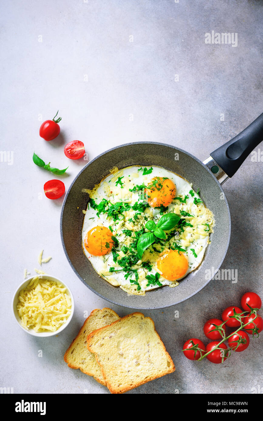 Fried eggs in pan, herbs, spices, cherry tomatoes, cheese toasts, basil for breakfast on grey concrete stone background. Copy space, top view, flat la Stock Photo