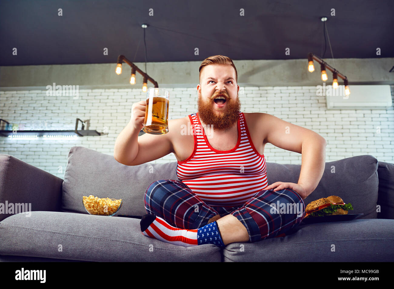 Thick funny man eating a burger sitting on the couch, watching TV at