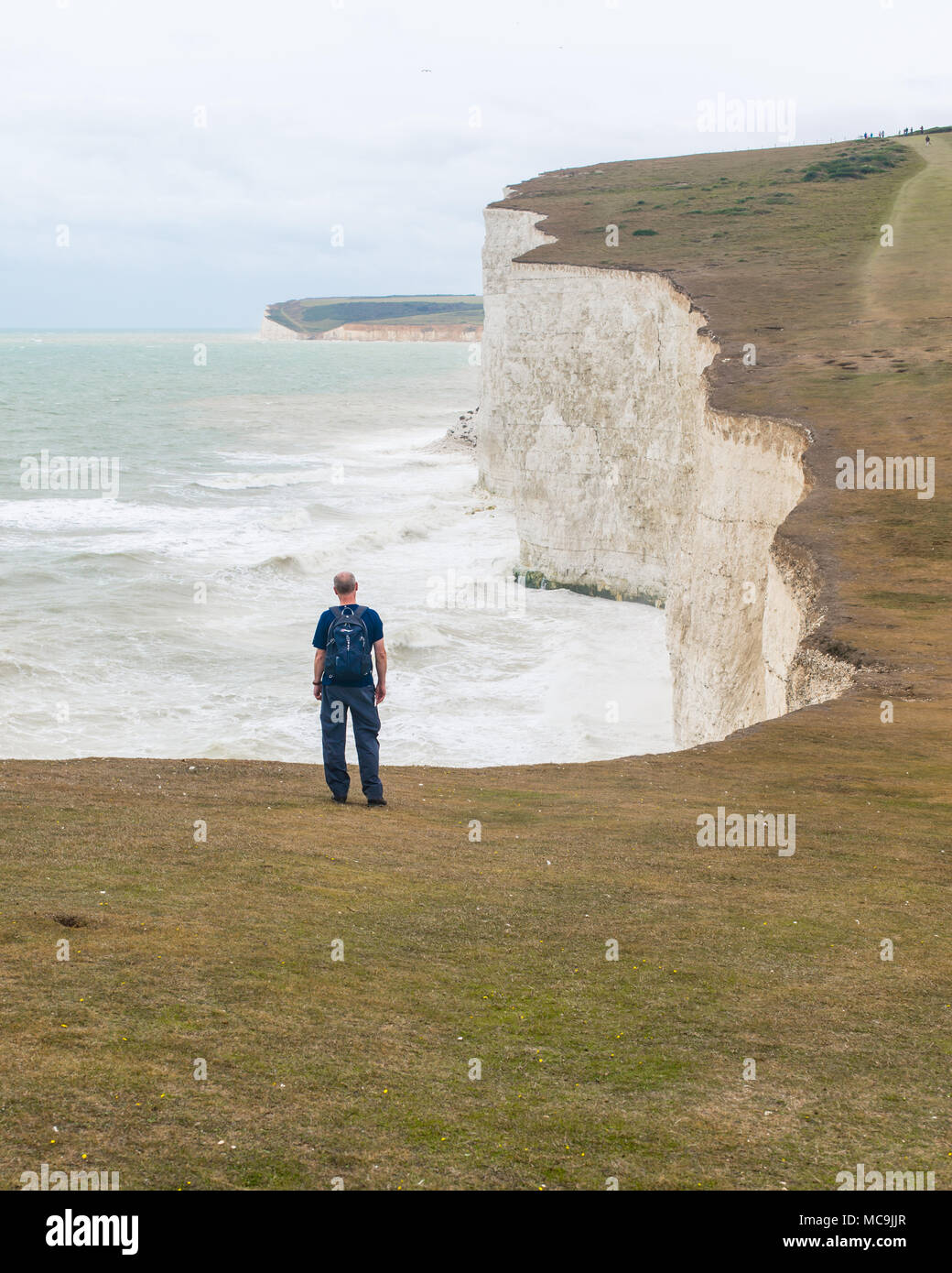 Male hiker wearing backpack from behind admiring the white cliffs of Seven Sisters facing rough sea and waves. Seven sisters, Seaford, UK Stock Photo