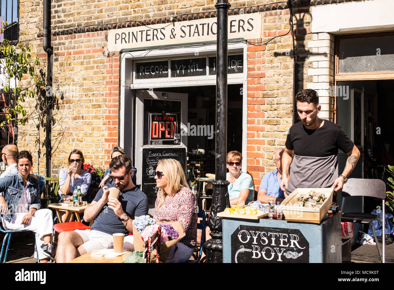 People drinking and eating al fresco on outside tables in front of hipster wine shop café-bar called Printers & Stationers. Bethnal Green, East London Stock Photo