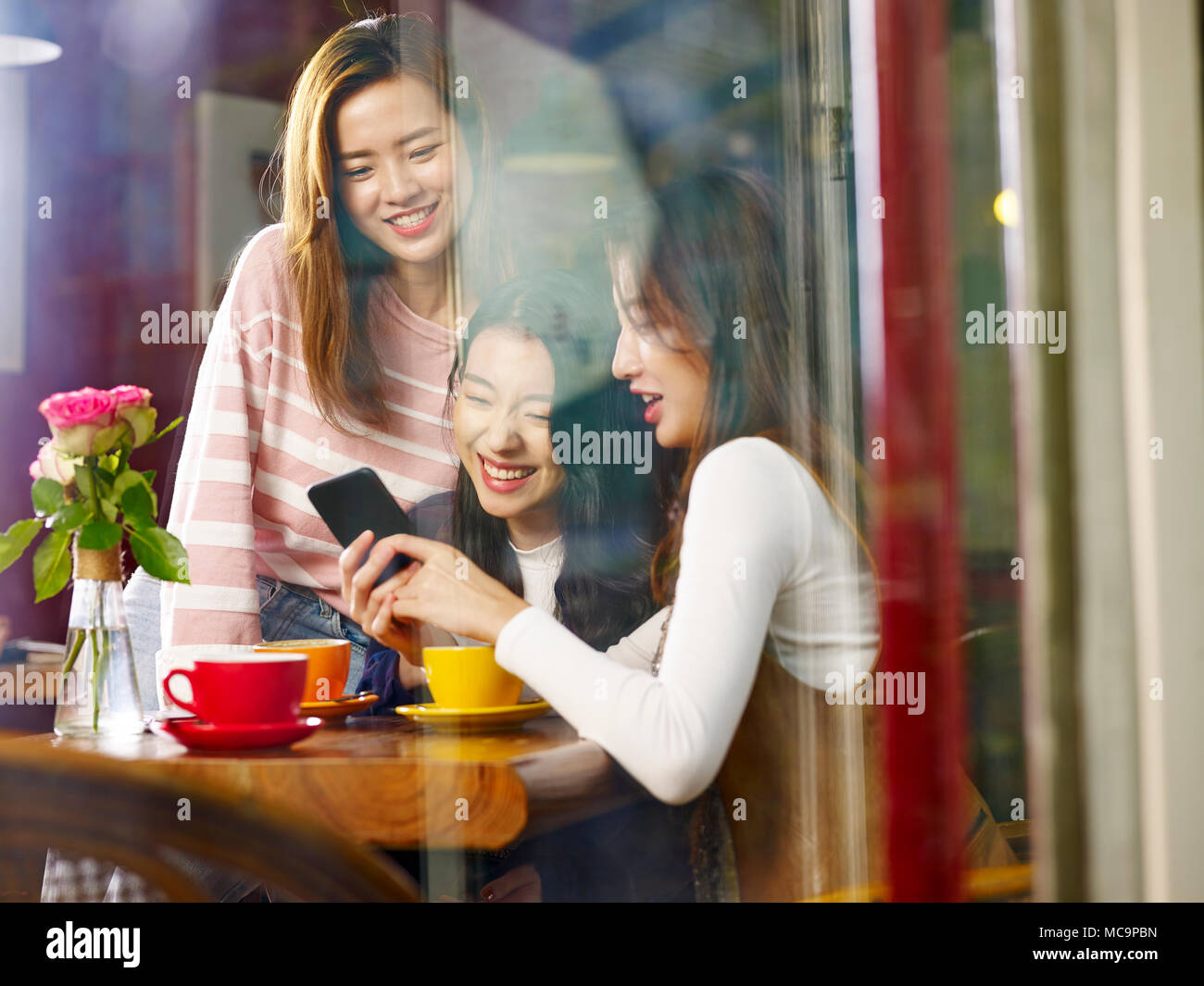 three happy beautiful young asian women sitting at table chatting talking playing with cellphone in coffee shop or tea house, shot through window glas Stock Photo