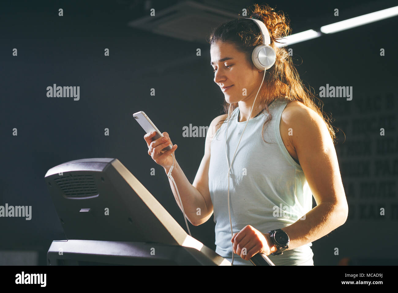 Theme sport and music. A beautiful Caucasian woman running in gym on  treadmill. On head big white headphones, the girl listens to music during a  cardio workout for weight loss and uses