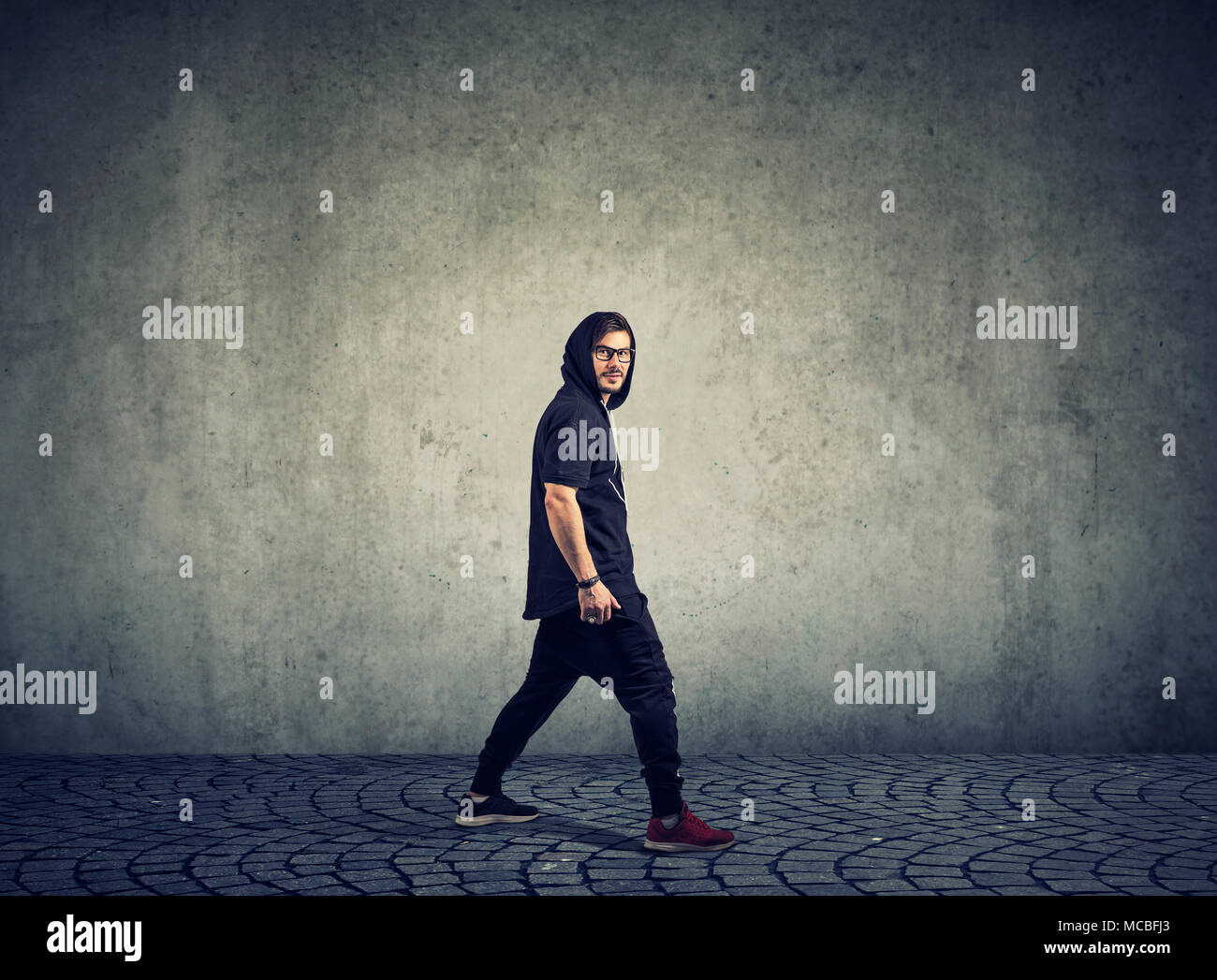 Side view of stylish hip-hop dancer in glasses walking in sportswear looking at camera. Stock Photo