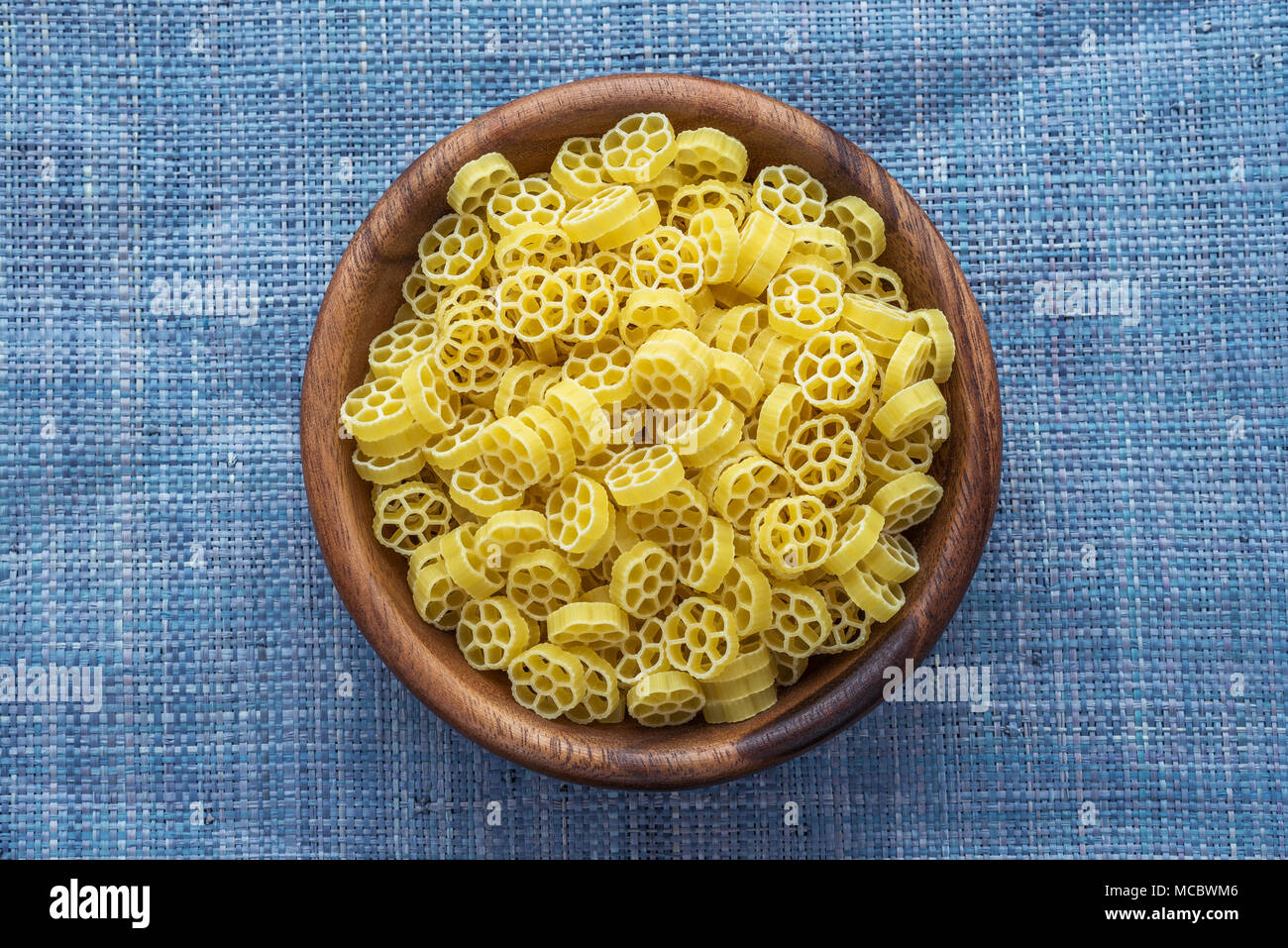 Macaroni ruote pasta in a wooden bowl on a blue knitted background in the center. Close-up with the top. Stock Photo