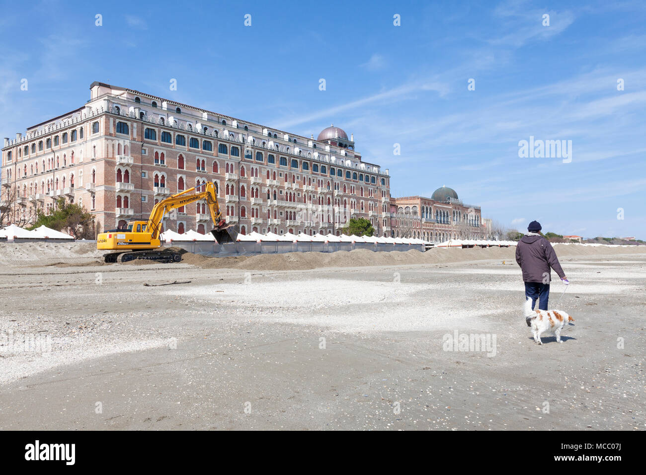 Man walking his dog on the beach, Lido di Venezia, Venice, Veneto, Italy passing the Excelsior Hotel and an excavator preparing the beach for the summ Stock Photo