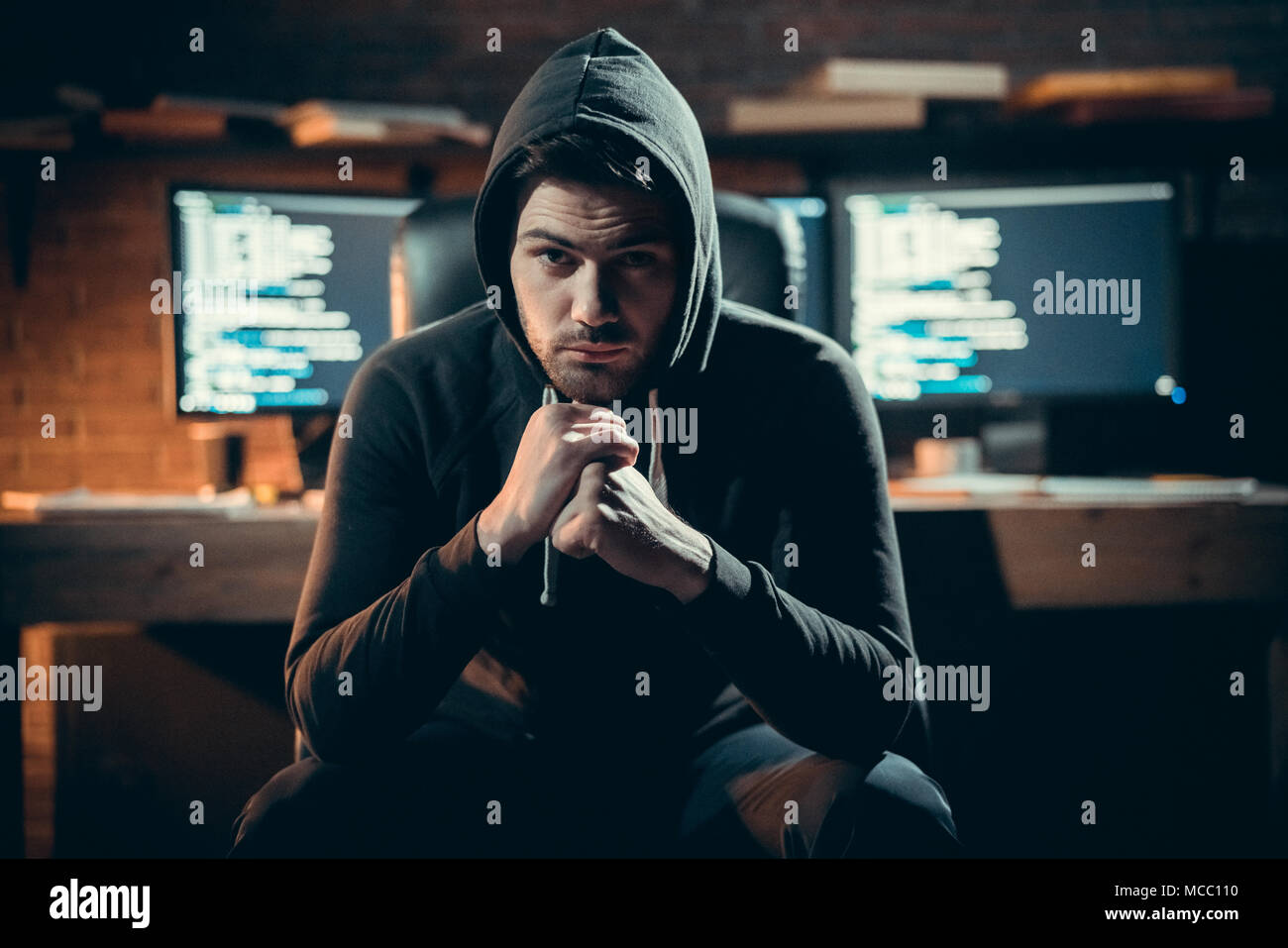 Portrait of young dangerous hooded hacker in black hoodie looking at camera posing with code on monitors at background, stealing information, cyber se Stock Photo