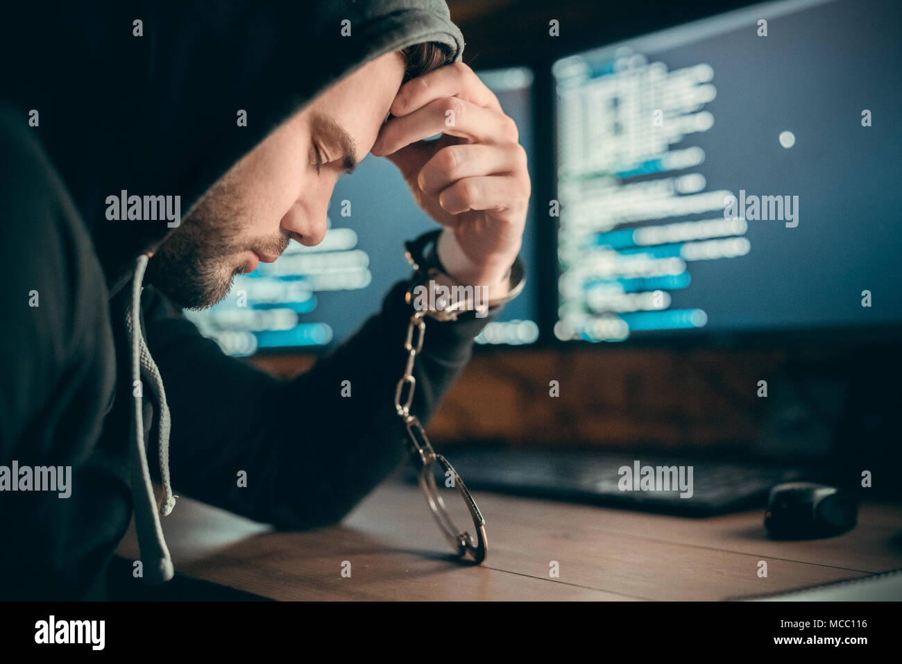 Arrested upset hacker or sad internet criminal got caught to be imprisoned sitting with hand in handcuffs on computer code background, cyber crime law Stock Photo