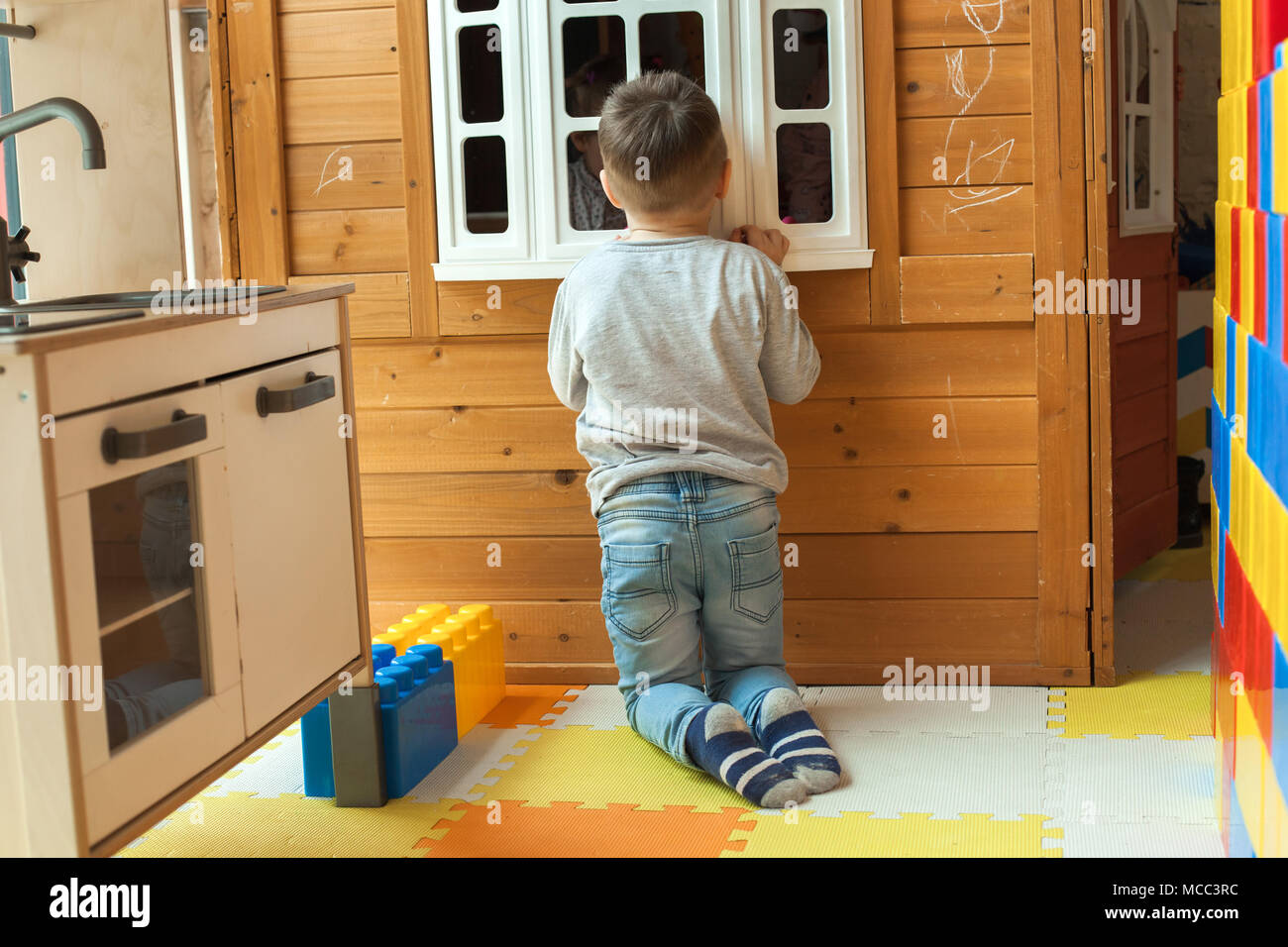 the boy is 4 years old, the blond plays on the playground indoors, peeps out the window of the toy wooden house Stock Photo
