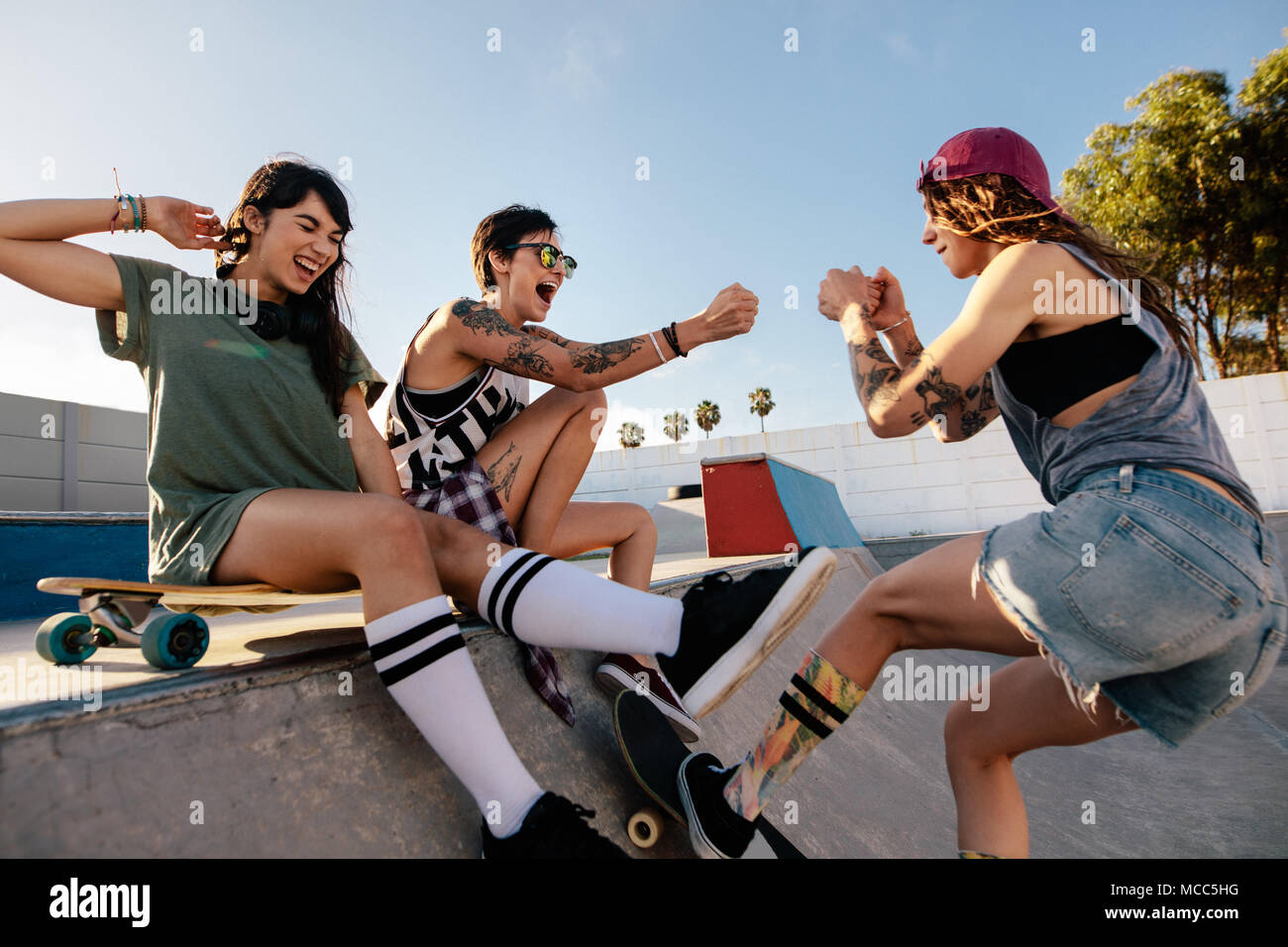 Three young women having a great time at skate park. Young female skateboarding with friends sitting on ramp. Stock Photo