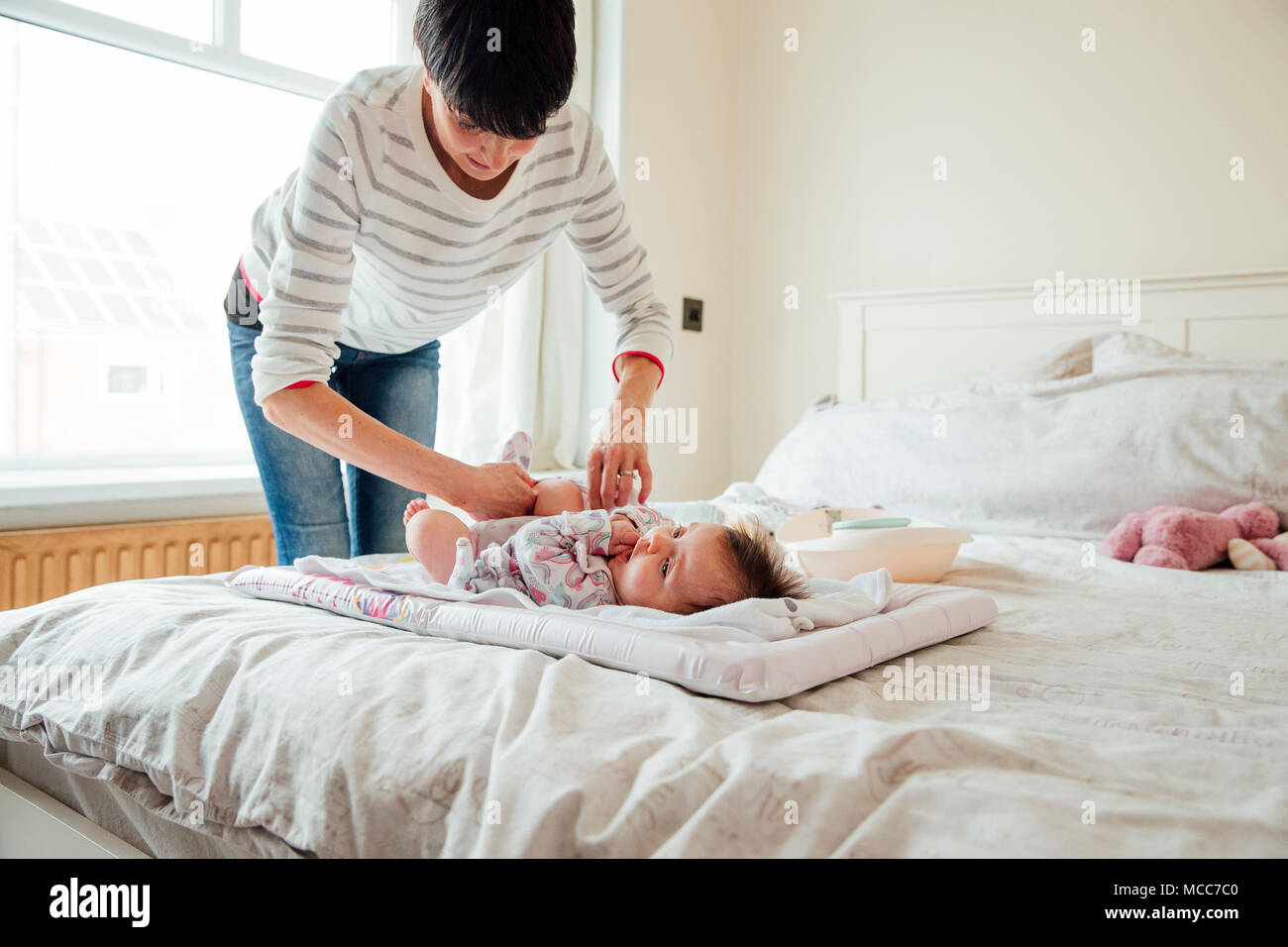 Baby has had her nappy changed by her mother on the double bed. The mother is fastening the baby grow. Stock Photo