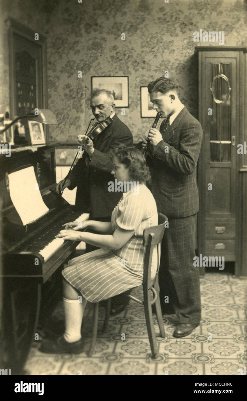 Historical picture @ 1900, showing a family members playing music together at home. The girl is sitting playing the piano, while her brother standing beside her plays the flute and next to him,the father the violin. Stock Photo