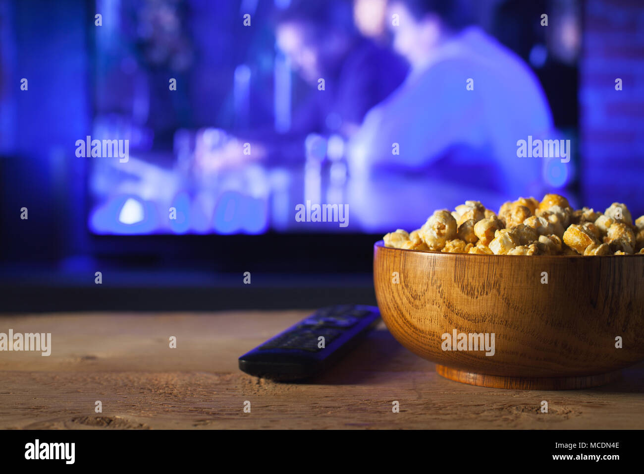 A wooden bowl of popcorn and the TV remote in the background the TV works. Evening cozy watching a movie or TV series at home. Stock Photo