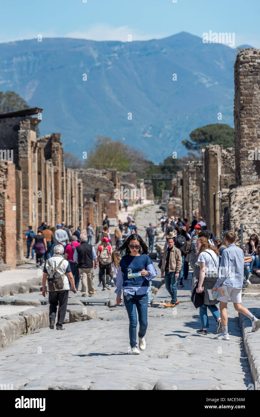 Female / woman tourist walking down center of street in Pompeii looking at map; tourists, mountain and ruins behind her. Stock Photo