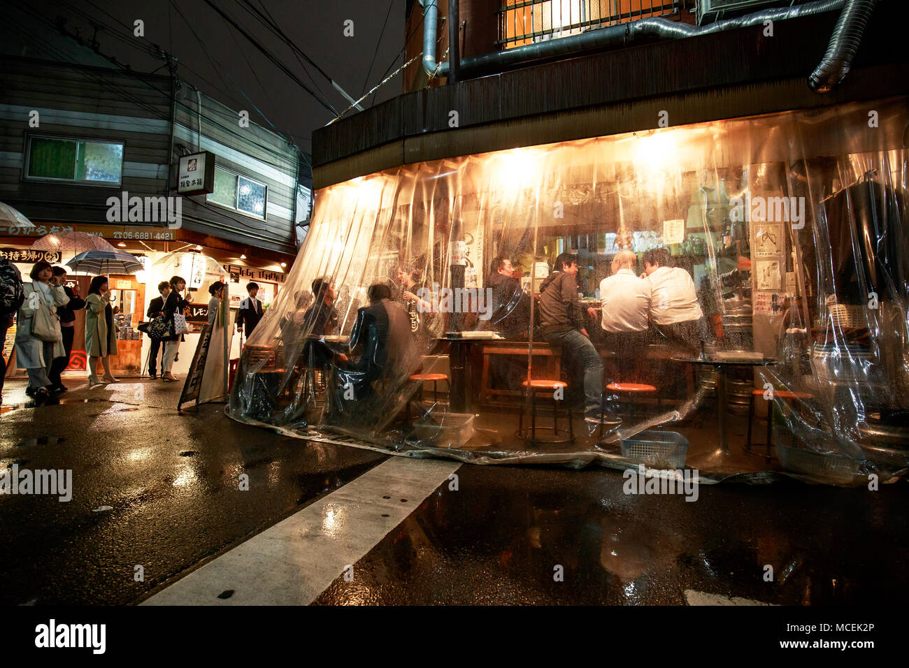 Osaka, Japan - April 14, 2018: Customers drink in a corner bar sheltered from the wind and rain by a clear plastic curtain Stock Photo