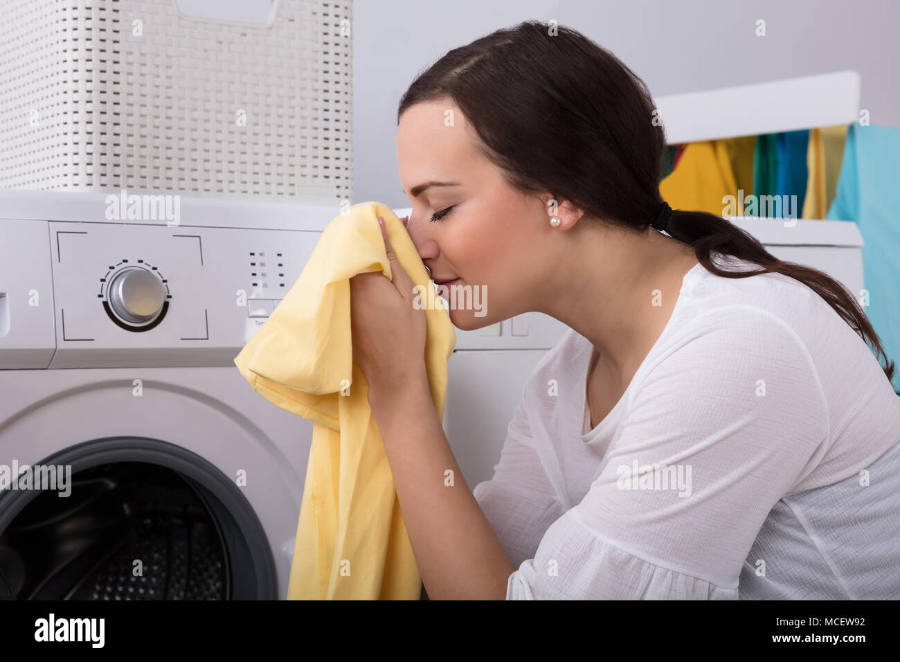 Side View Of A Young Woman Smelling Cleaned Yellow Cloth Near Washing Machine Stock Photo