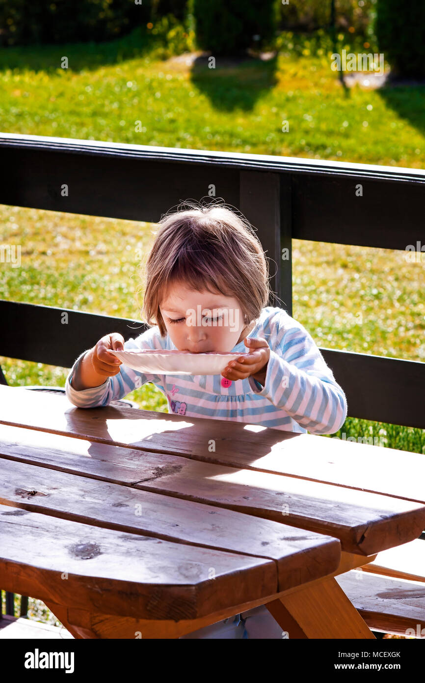 Little girl  lapping her milk soup directly from plate , outdoors. Stock Photo