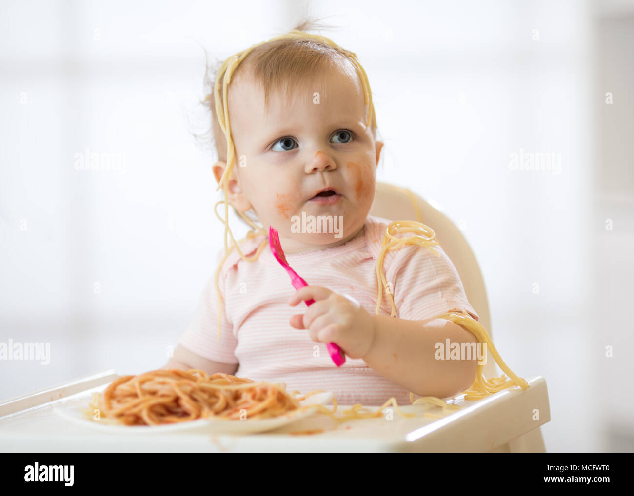 Funny child eating noodle. Grimy kid eats spaghetti with fork sitting on table at home Stock Photo