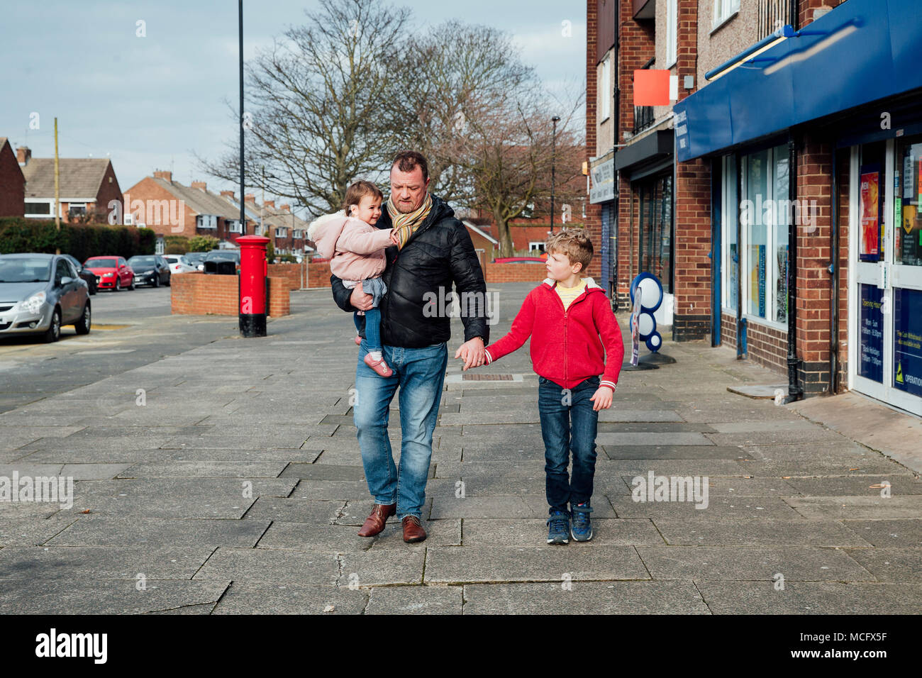 Mature man is taking his children to the local shop with him. He is holding his sons hand and carrying his baby daughter on his hip. Stock Photo