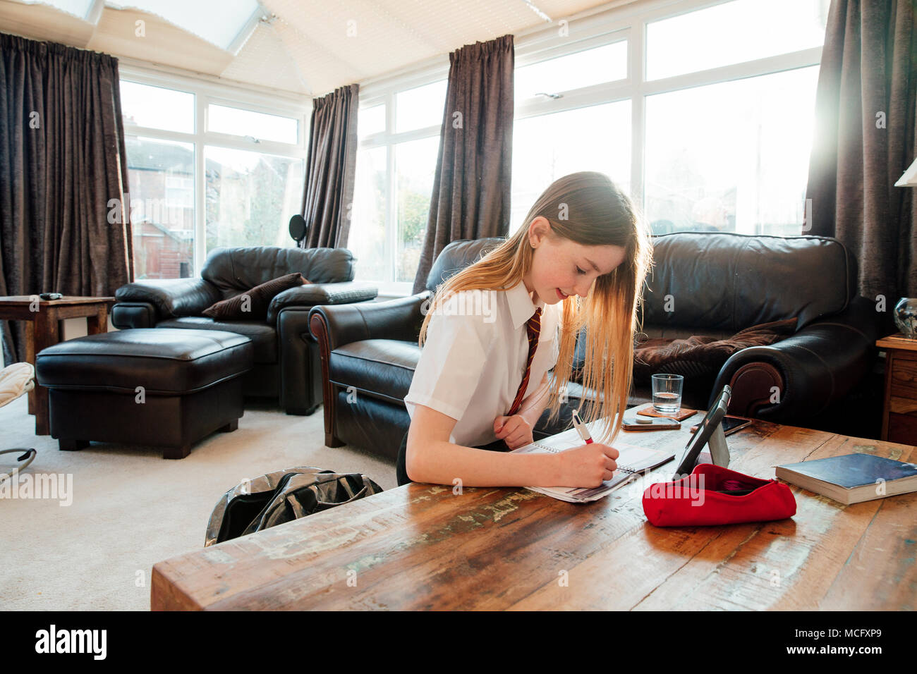 Girl is doing homework in the living room if her home. She is writing in a book, using a digital tablet to study. Stock Photo